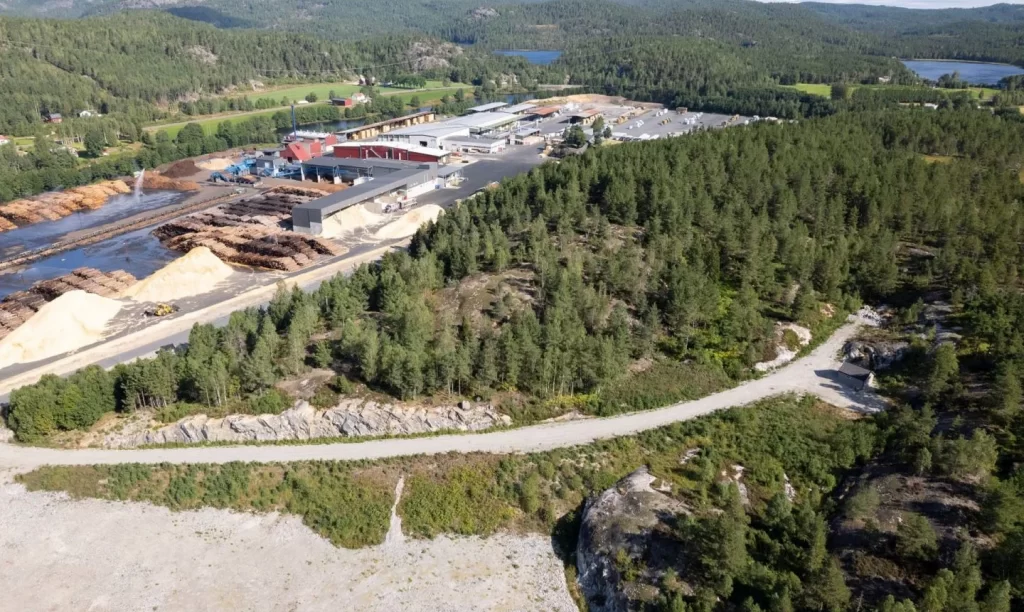 Fabrikken vil bli oppført på tomten til høyre i bildet som er regulert til dette formålet. Til venstre er Nidarå sagbruket som er eid og driftet av Bergene Holm AS. Sidestrømmer fra sagbruket vil kunne benyttes i produksjonen.